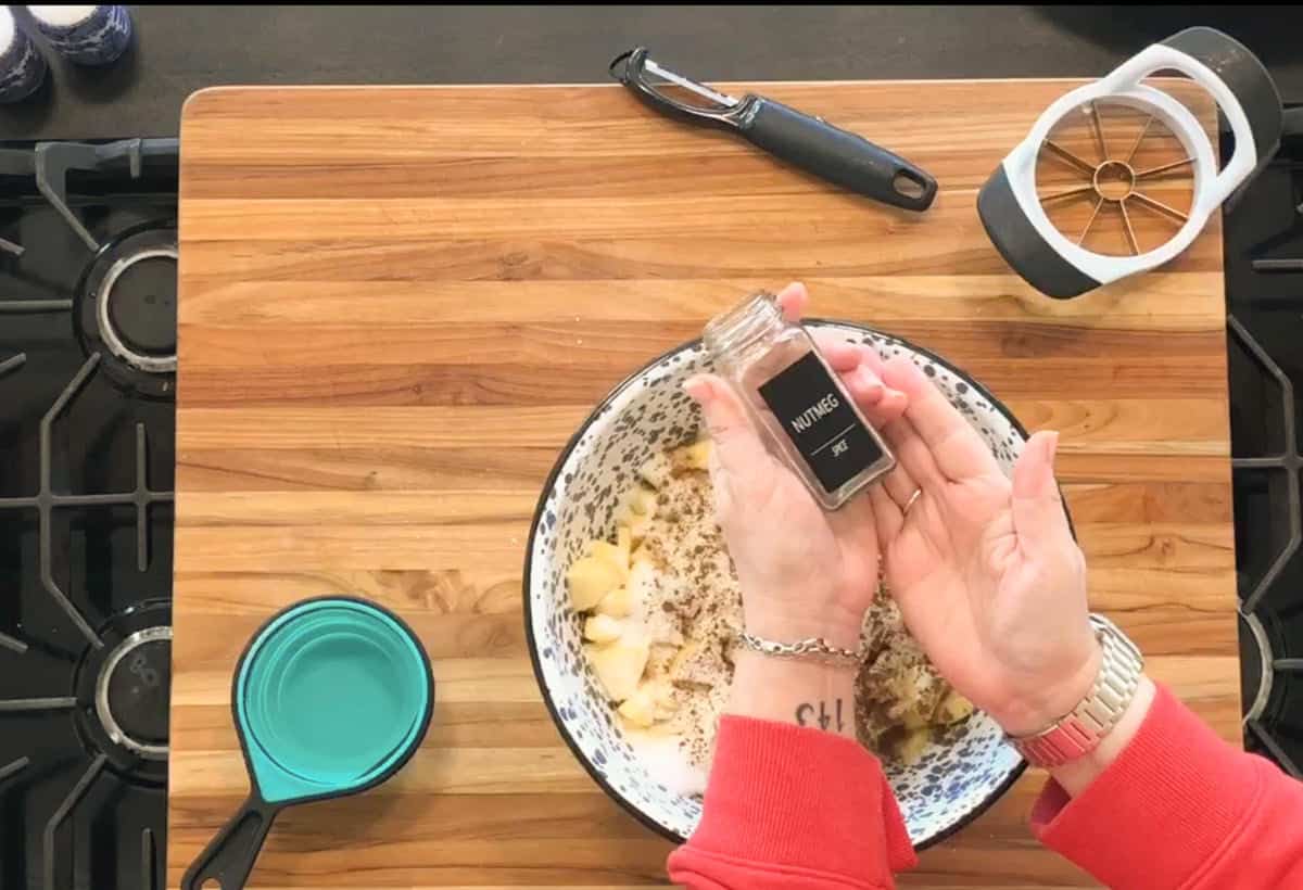 A person sprinkles ground nutmeg from a spice jar into a bowl of chopped apples on a wooden cutting board. A blue measuring cup, apple slicer, and peeler are nearby.