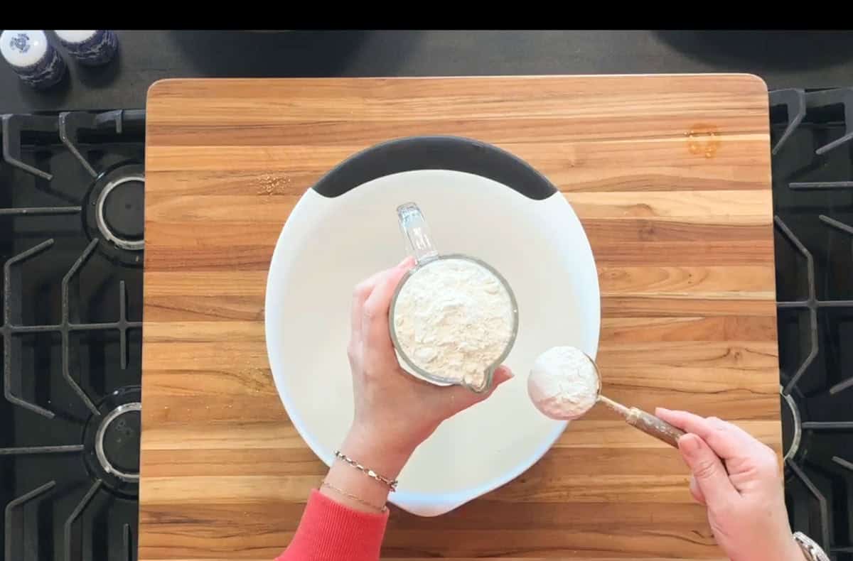 A person holds a measuring cup filled with flour over a large white mixing bowl on a wooden cutting board, using a spoon to level off the flour. A stove is visible on either side of the cutting board.