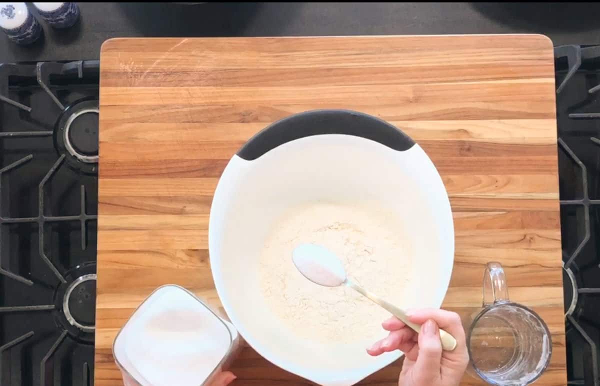 A hand holds a spoonful of sugar above a white mixing bowl filled with flour on a wooden cutting board. A container of sugar and a glass measuring cup with water are nearby. The scene is on a stovetop.