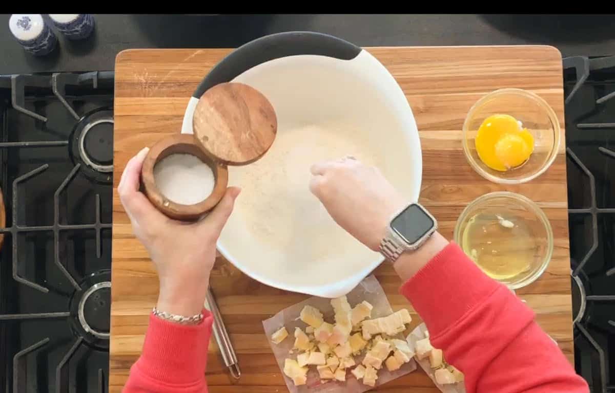 A person wearing a red sleeve and a watch adds salt to a large white mixing bowl with flour. On the cutting board are chopped butter, a bowl with an egg yolk, and a bowl with egg whites. The scene is on a stove.