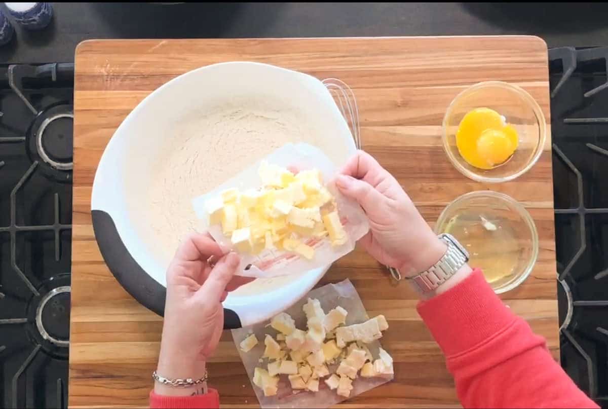 A person in a red sleeve adds cubed butter into a white mixing bowl filled with flour. Two small bowls with egg yolks and egg whites are on the wooden countertop next to the bowl.