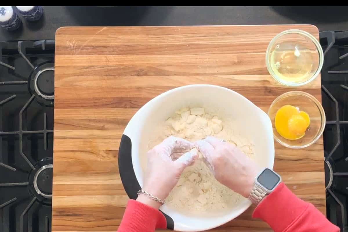 A person wearing a red sleeve and watch mixes dough in a white bowl on a wooden cutting board. Two bowls beside the main bowl contain eggs, and the scene is set on a black stovetop.