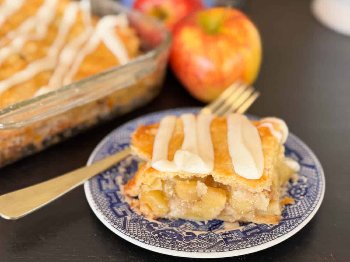 A slice of apple pie with icing on top sits on a blue and white plate with a fork. Behind it, a glass dish with the rest of the pie and whole apples are visible.