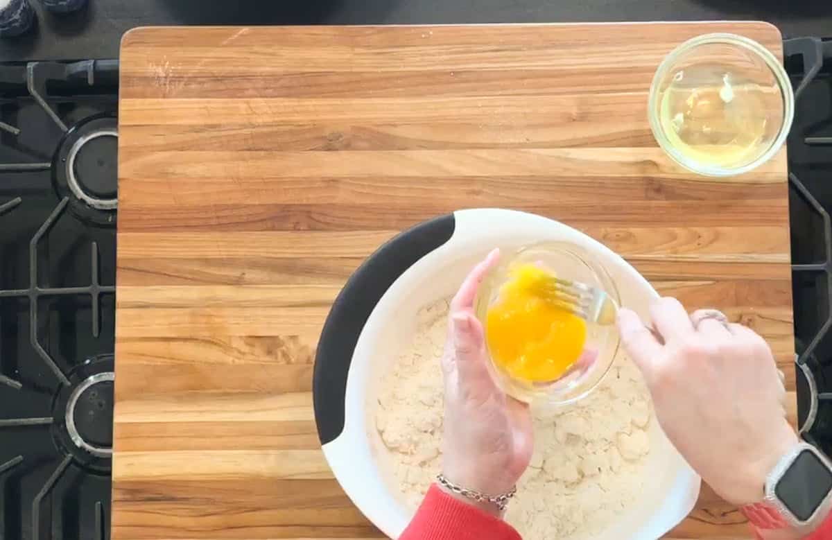 A person holds a small bowl with beaten eggs over a mixing bowl containing flour on a wooden cutting board. An unbroken egg is in a separate small bowl nearby.