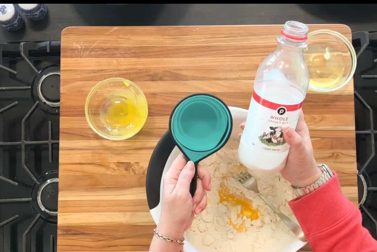 A person holds a bottle of whole milk and a measuring cup above a bowl of flour and cracked eggs on a wooden cutting board. Two small bowls with eggs are nearby on the counter.