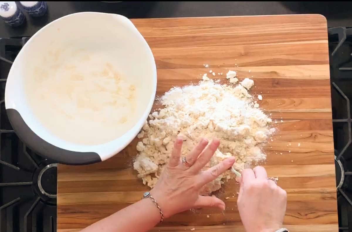 Two hands mix flour and dough on a wooden surface next to an empty mixing bowl; the dough appears partially combined with some flour still visible.