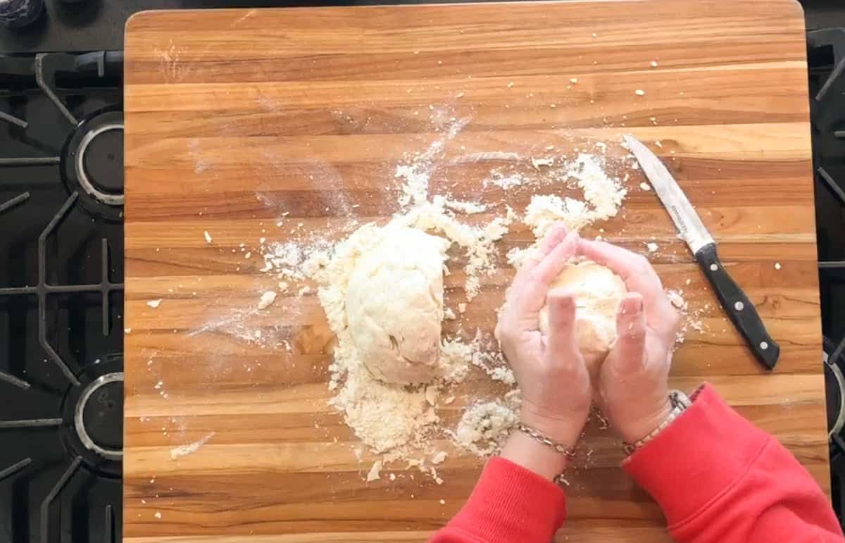 Hands in a red sleeve shaping dough on a wooden cutting board. A knife is placed nearby, and flour is scattered across the board. The scene is on a stovetop.