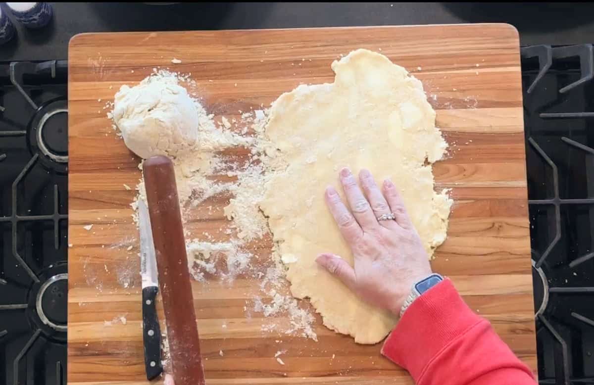 A person in a red sleeve presses dough flat with their left hand on a wooden cutting board. A ball of dough, a rolling pin, and a knife are visible, with flour scattered across the board.