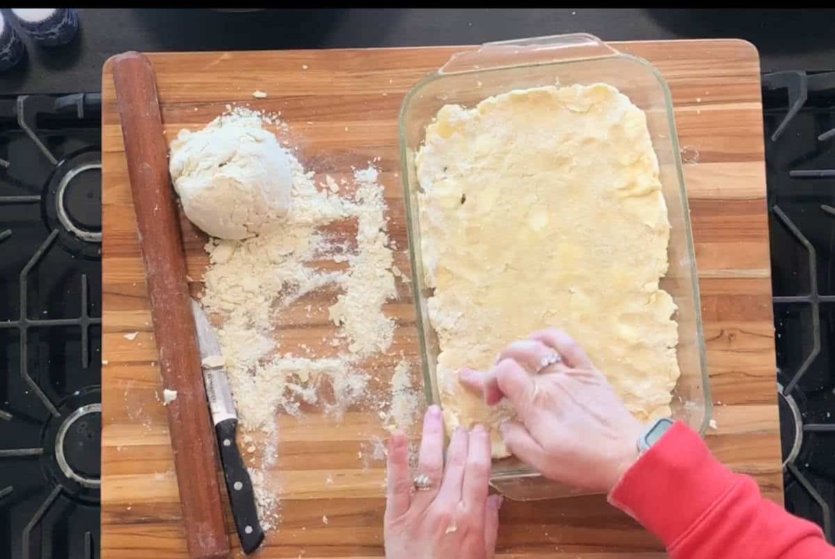 A person presses pie dough into a glass baking dish on a wooden surface. A rolling pin, dough ball, and knife dusted with flour are nearby. The baking is being done on a stovetop.