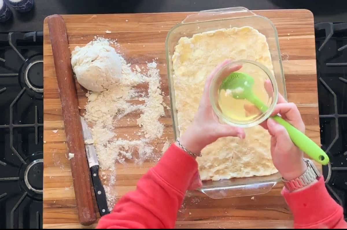 A person in a red sleeve holds a bowl with a whisked egg over a rectangular baking dish lined with dough. A rolling pin, knife, flour, and extra dough are on a wooden cutting board.