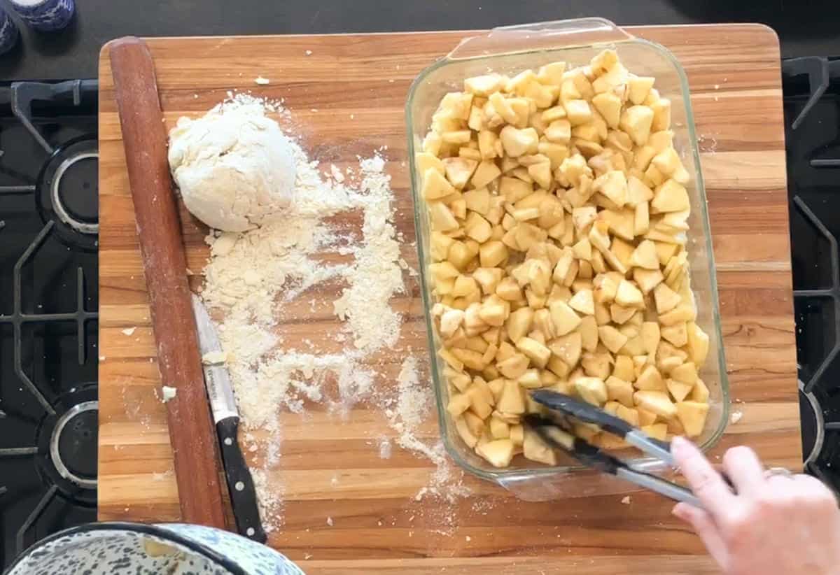 A hand uses tongs to arrange chopped apples in a glass dish on a wooden cutting board. Nearby are a ball of dough, a rolling pin, a knife, and some spilled flour.
