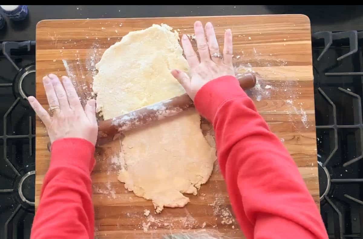 A person wearing a red sweatshirt uses a wooden rolling pin to roll out dough on a floured wooden cutting board placed on a stove.