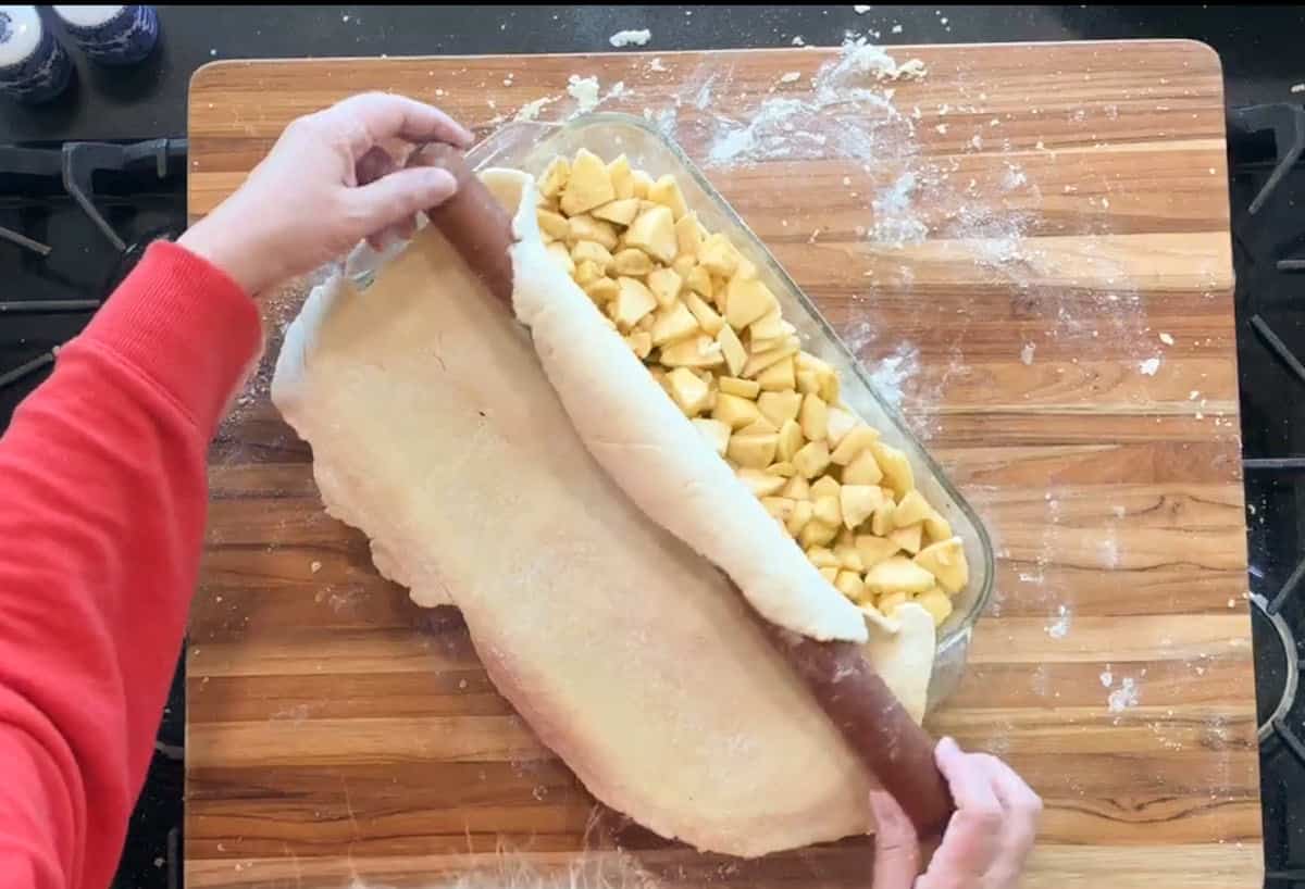 Person in a red sleeve rolls out dough over a glass baking dish filled with chopped apples on a wooden surface, preparing to cover the filling for baking. Flour is scattered on the board.