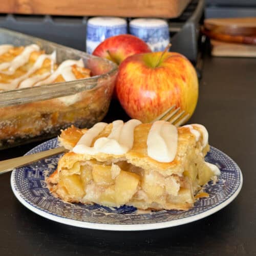 A slice of glazed apple pie with icing sits on a blue and white plate in front of a glass baking dish and two whole apples on a dark surface. A fork rests on the plate beside the pie.