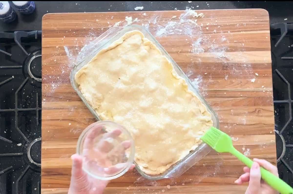 A person holds a small glass bowl and uses a green pastry brush to coat the top crust of an unbaked pie in a rectangular glass dish on a wooden surface dusted with flour.