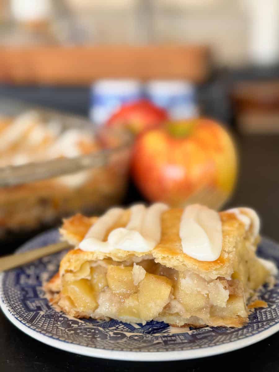 A slice of apple pie with a golden-brown crust and white icing sits on a blue-patterned plate, with a blurred whole pie and a red apple in the background.