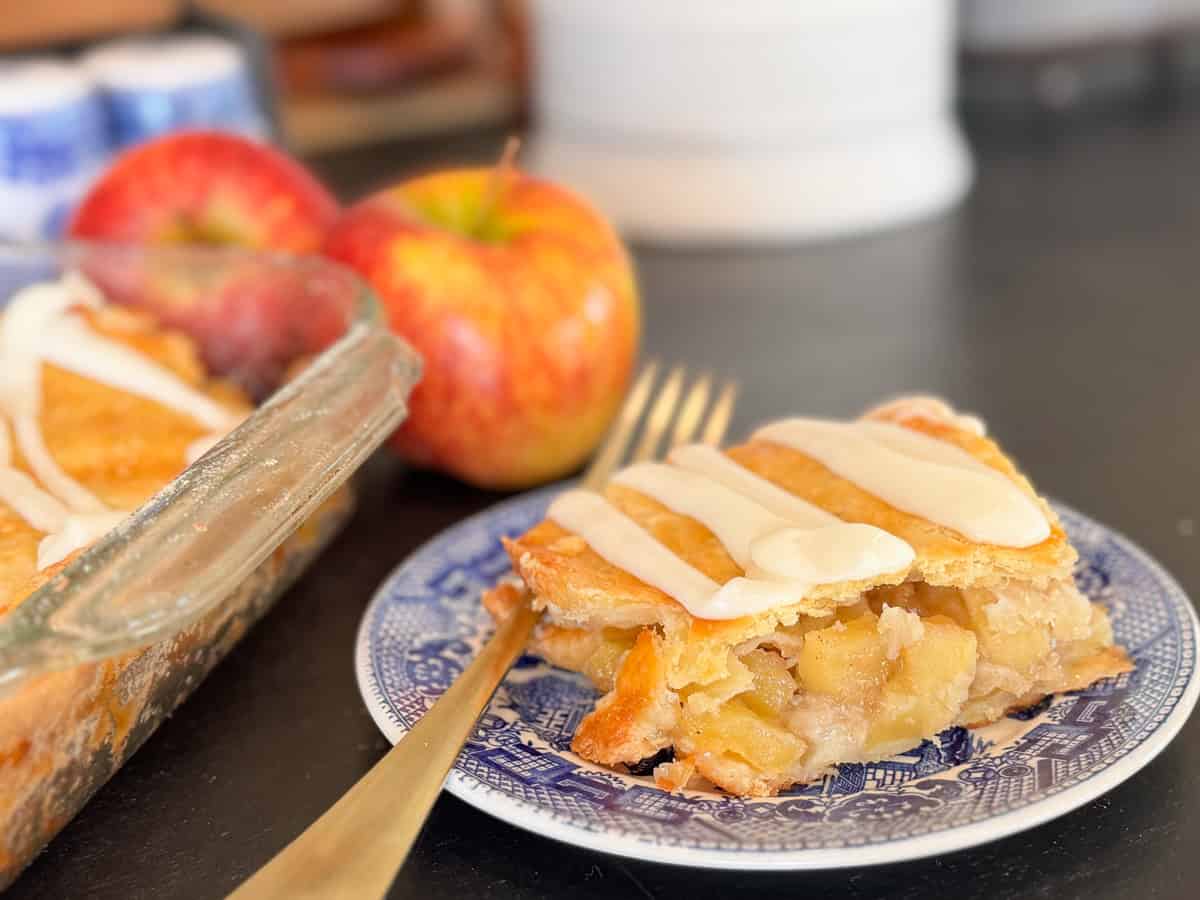 A slice of apple pie with icing sits on a blue patterned plate with a fork. The pie has a flaky crust and visible apple filling. In the background are whole apples and a baking dish with more pie.