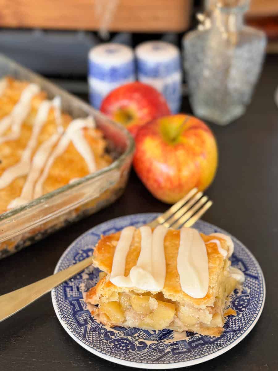 A slice of apple pie with white icing sits on a blue and white plate, next to a gold fork. Behind it are two red apples, a glass baking dish of pie, salt and pepper shakers, and a glass jar.