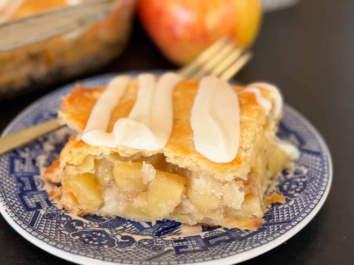 A slice of apple pie with a golden-brown crust and visible apple filling, topped with three lines of white icing, sits on a blue and white patterned plate. An apple and a fork are in the background.