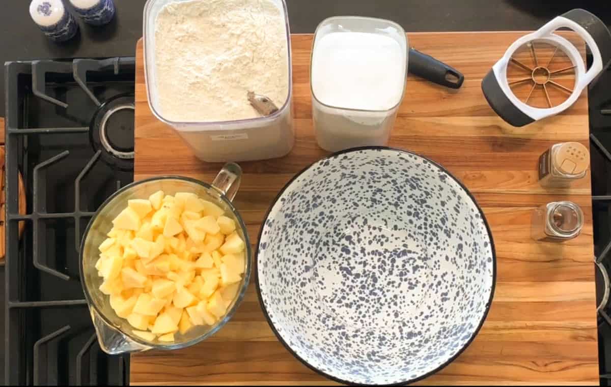 A wooden countertop with a speckled mixing bowl, a bowl of diced apples, containers of flour and sugar, an apple slicer, a peeler, and salt and pepper shakers arranged neatly on top.