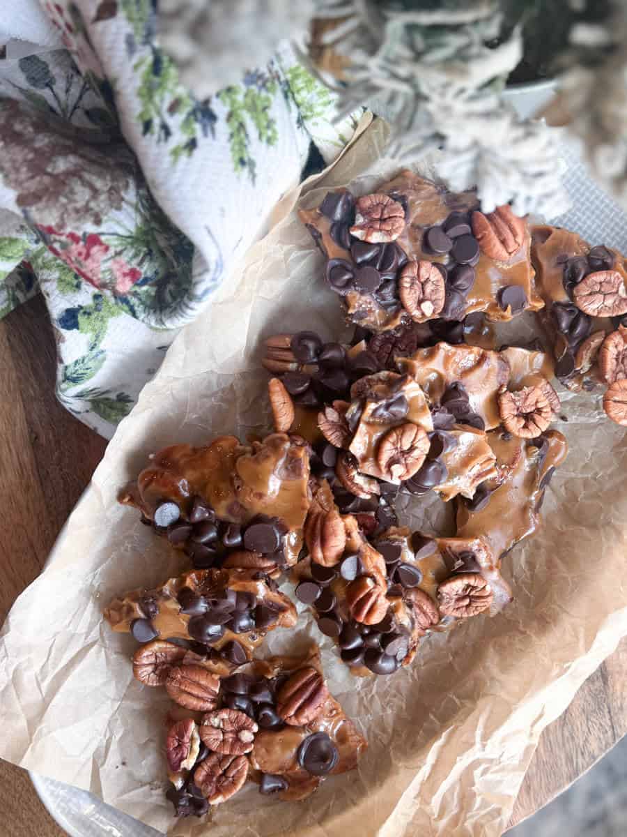 Homemade candy bark topped with pecan halves and chocolate chips, resting on a sheet of parchment paper. A holiday-themed towel and snowy artificial tree branches are partially visible in the background.