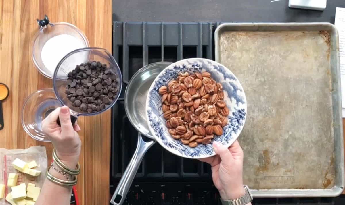 A person holds a bowl of pecans in one hand and a bowl of chocolate chips in the other over a stovetop, with a saucepan, baking sheet, butter, and other ingredients nearby on a wooden counter.