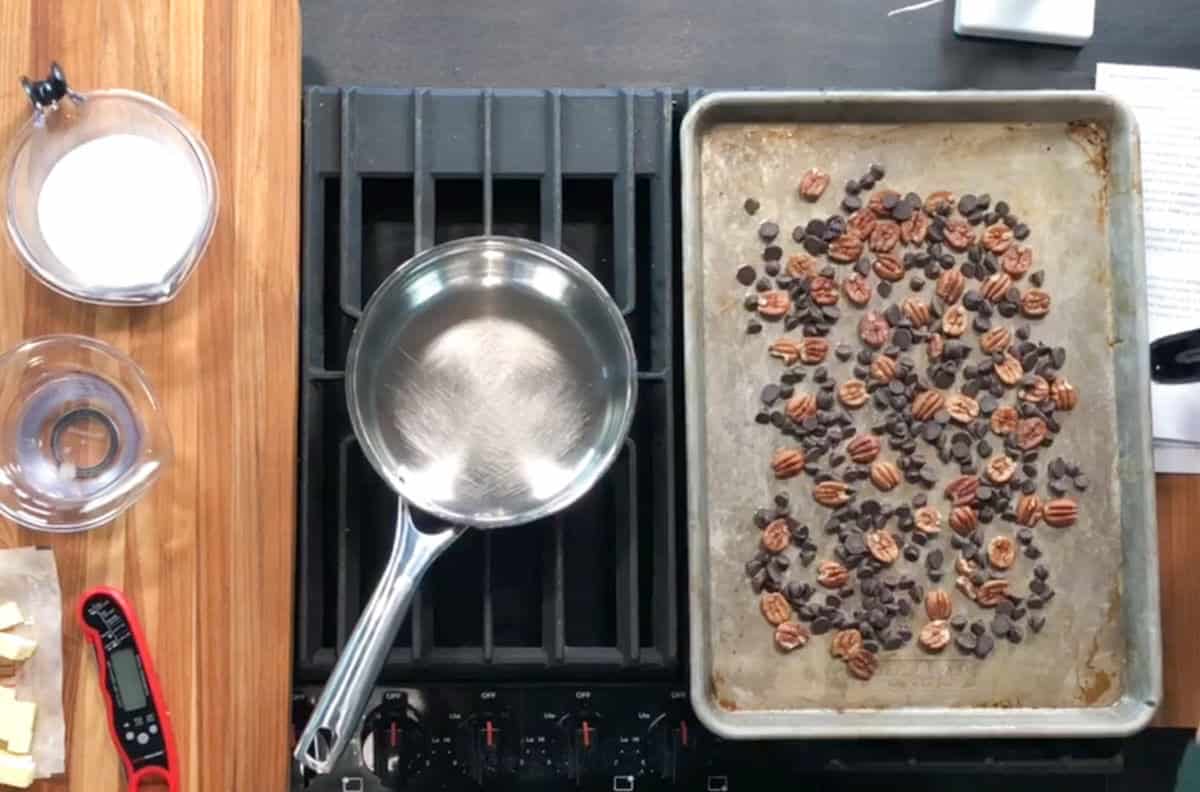 Overhead view of a stovetop with a metal saucepan, a baking sheet with scattered pecans and chocolate chips, and a wooden counter holding bowls of sugar, butter, a digital thermometer, and a paper with notes.