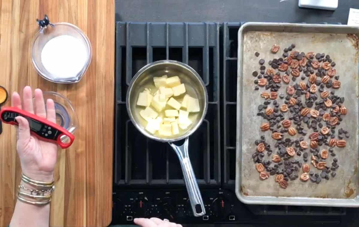 Overhead view of a stovetop with a saucepan of cubed butter, a baking sheet with chocolate chips and pecans, a hand holding a red thermometer, and measuring cups with ingredients on a wooden cutting board.
