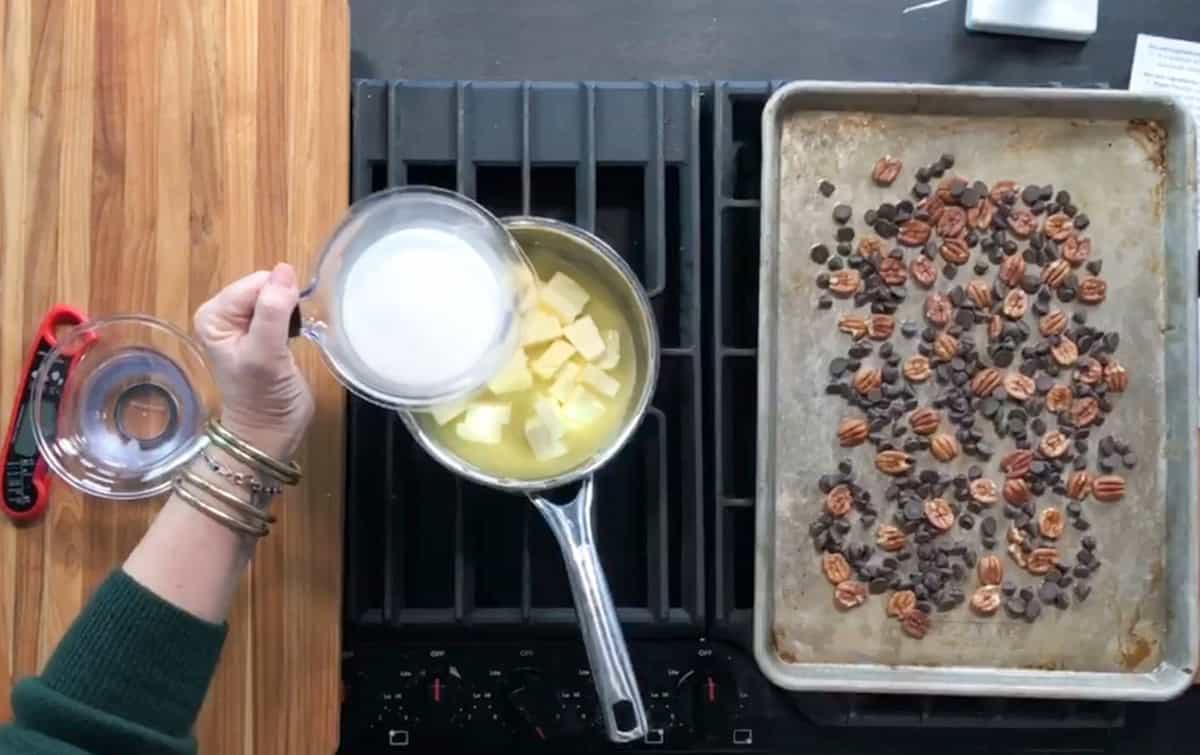 A person pours milk into a saucepan with melted butter on a stove. Next to the stove, a baking sheet with scattered pecans and chocolate chips sits on the counter. A measuring cup and lid are nearby on a wooden surface.