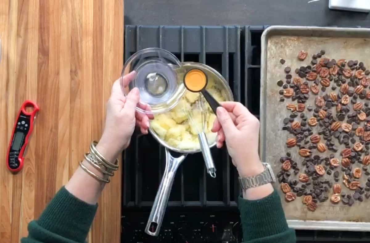A person adds a spoonful of liquid to a saucepan with chopped apples on a stove. A baking sheet with pecans and chocolate chips and a cooking thermometer are on the counter nearby.