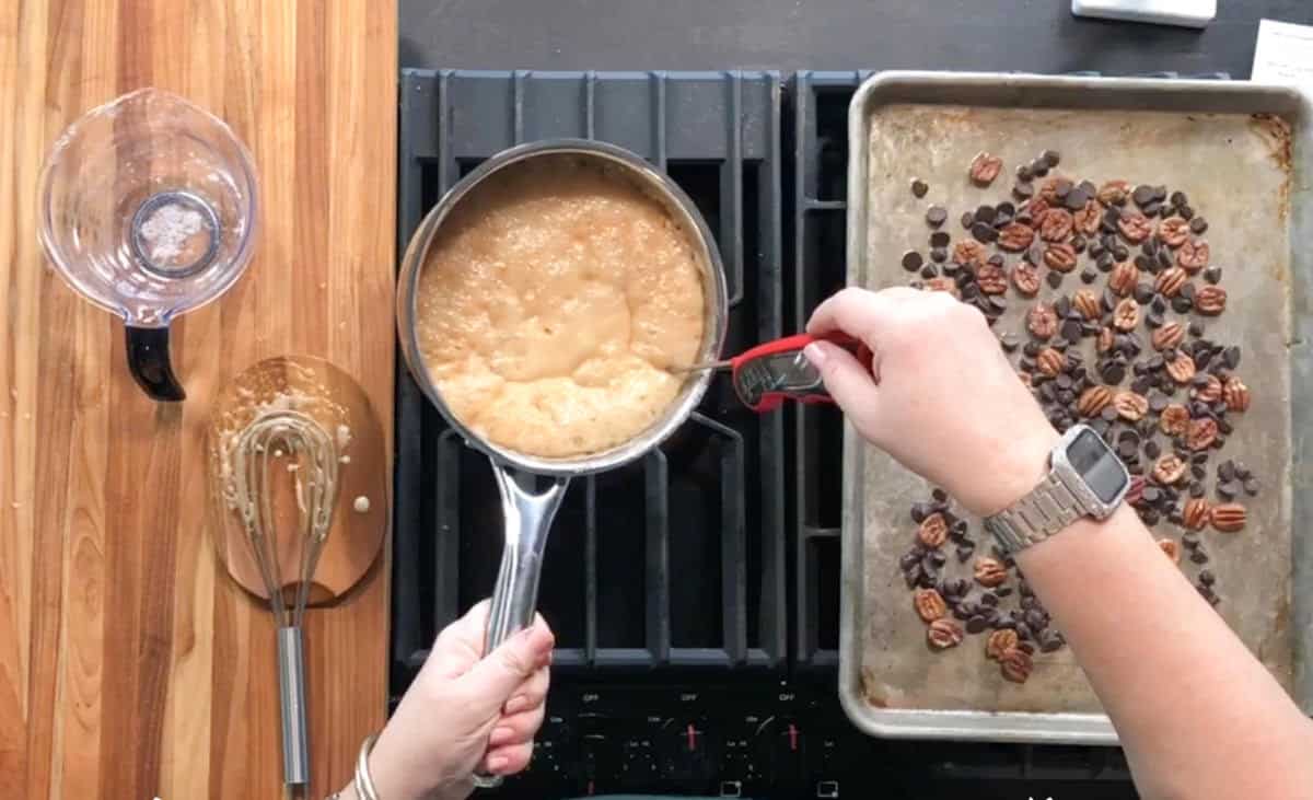 A person stirs bubbling caramel in a saucepan on a stove while holding a thermometer; beside the stove is a baking sheet with pecans and chocolate chips, a whisk, and a measuring cup.