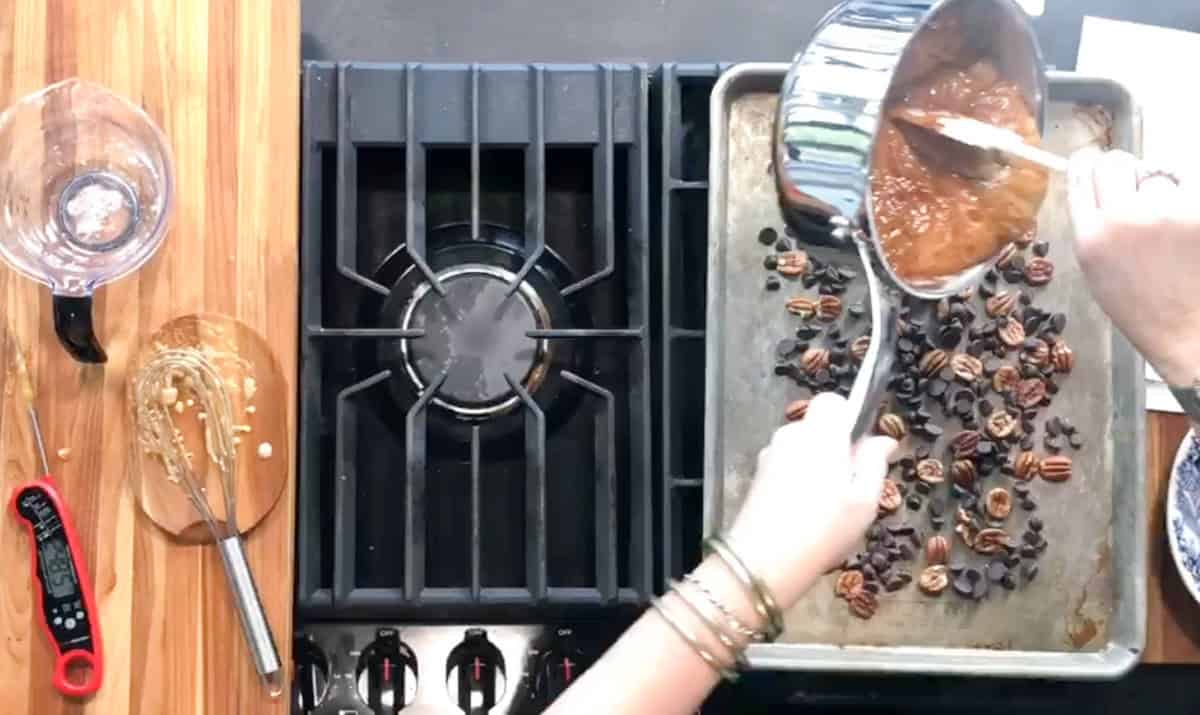 A person pours caramel from a saucepan onto a baking sheet lined with pecans and chocolate chips next to a stove, with a digital thermometer and whisk on the wooden counter.