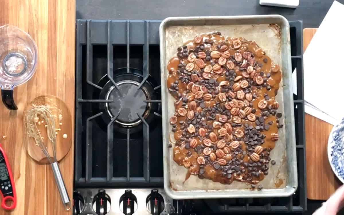 A baking sheet with a mixture of pecans, chocolate chips, and caramel sits on a stovetop. A whisk with caramel, a glass mug, a thermometer, and papers are on the wooden counter around the stove.