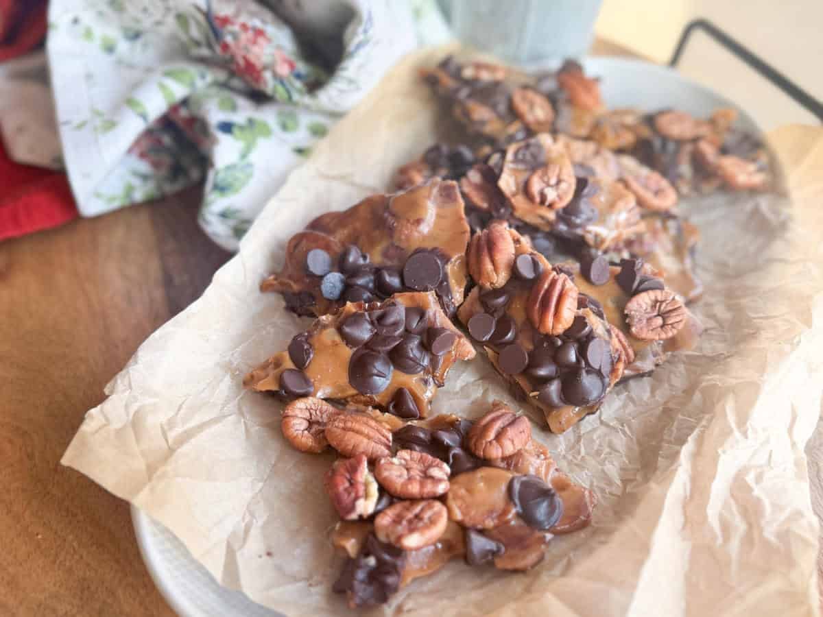 Broken pieces of toffee covered with pecans and chocolate chips are arranged on parchment paper atop a white plate, with a floral cloth in the background.