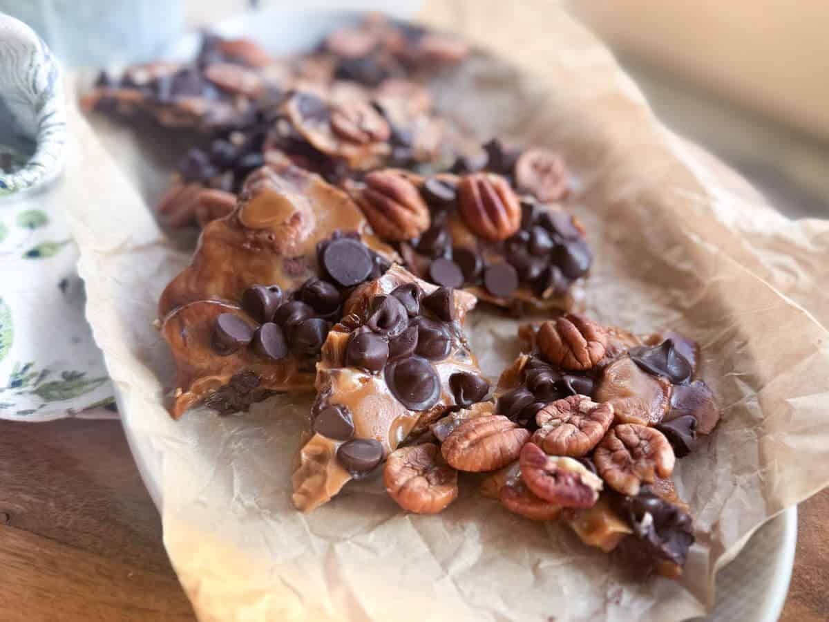 Pieces of homemade chocolate bark topped with pecans and chocolate chips are arranged on crumpled parchment paper on a wooden surface.