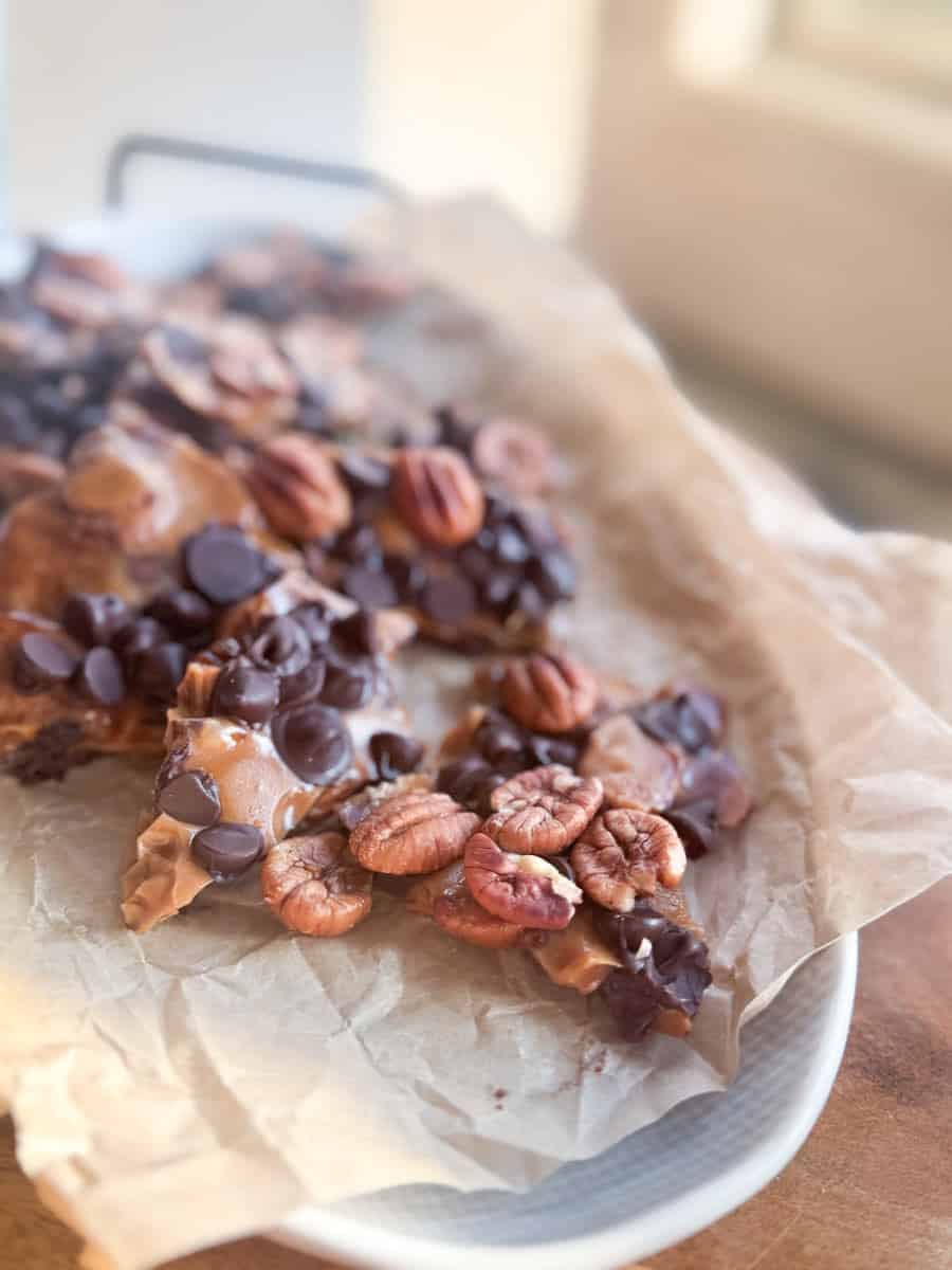Several pieces of chocolate bark topped with pecans and chocolate chips are arranged on a sheet of crumpled parchment paper atop a round white platter. The scene is softly lit by natural light.