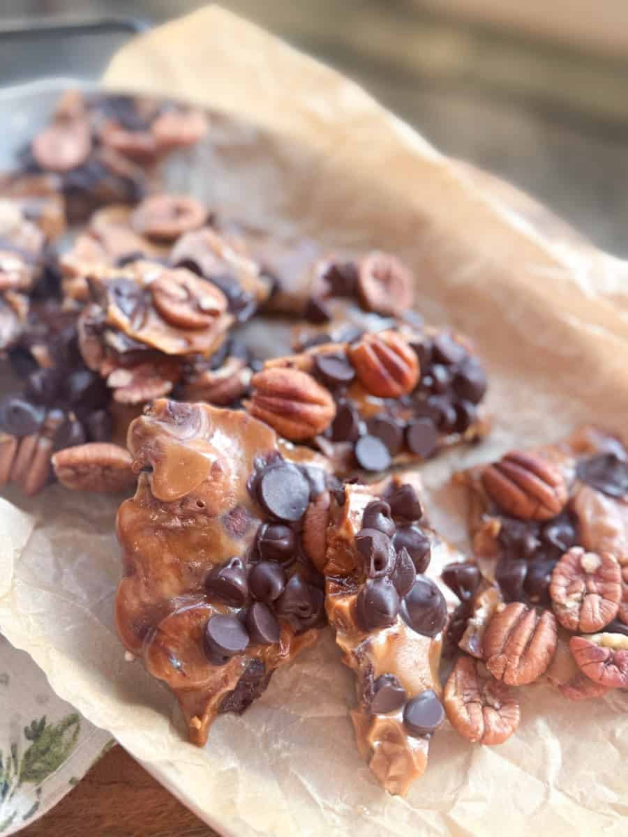 Pieces of nut brittle topped with whole pecans and chocolate chips are arranged on a sheet of parchment paper. The treats have a glossy surface and are set on a wooden surface in natural light.