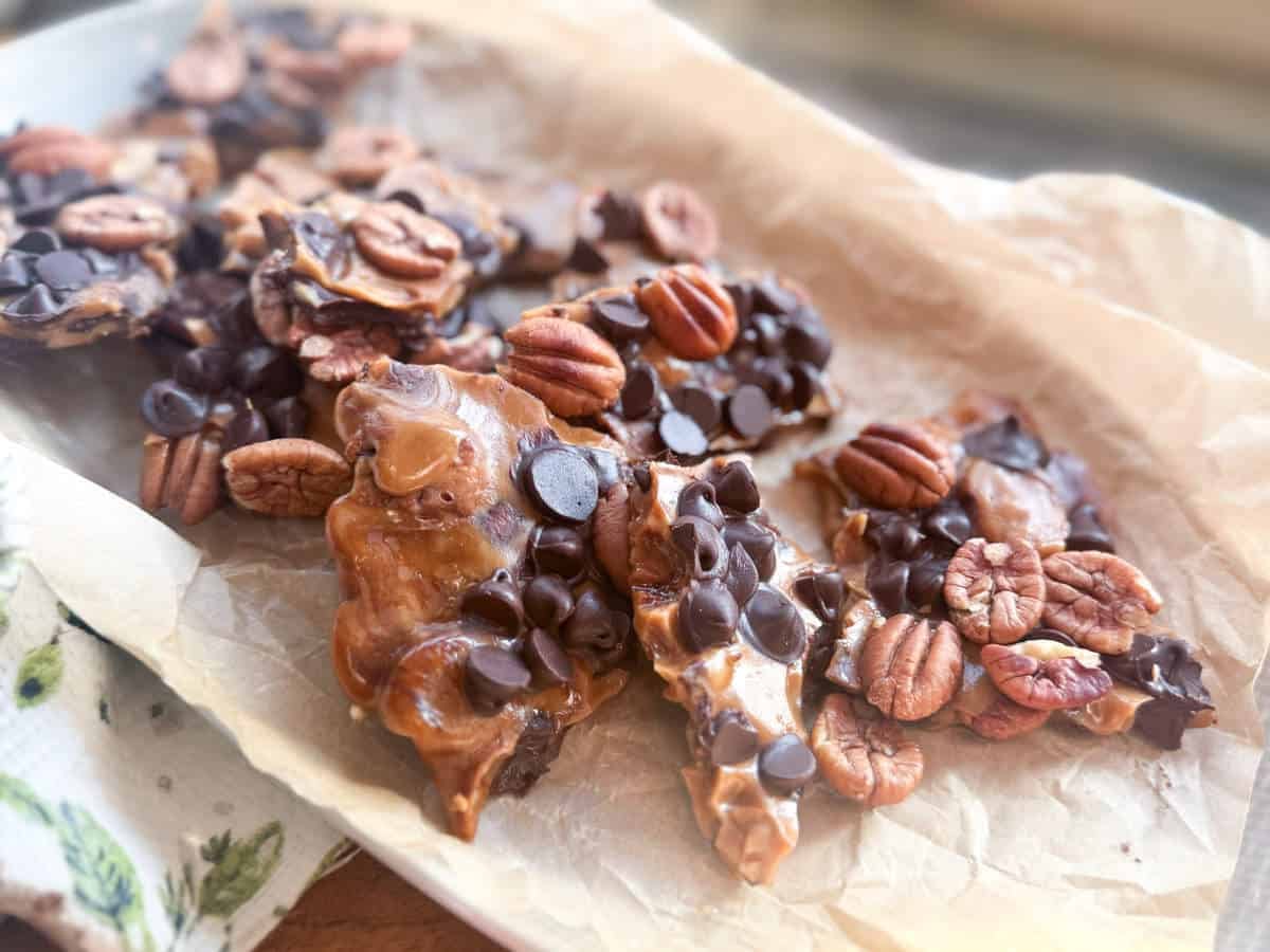 A close-up of chocolate pecan toffee bark pieces, topped with whole pecans and chocolate chips, arranged on crumpled parchment paper.