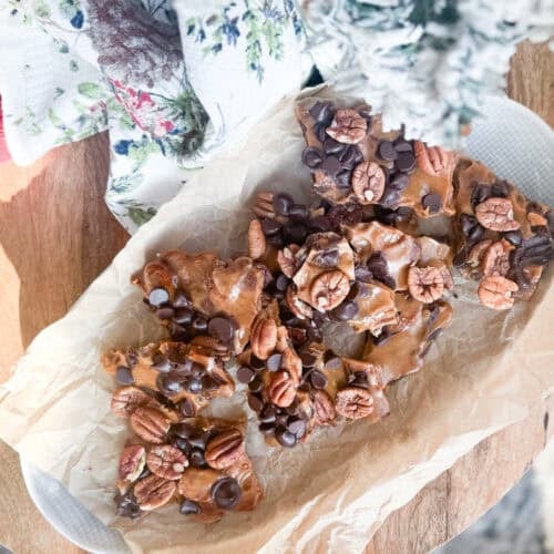 Parchment paper on a plate holds pieces of chocolate and pecan bark, topped with whole pecans and chocolate chips. A floral-patterned cloth and a snowy artificial tree are partially visible nearby.