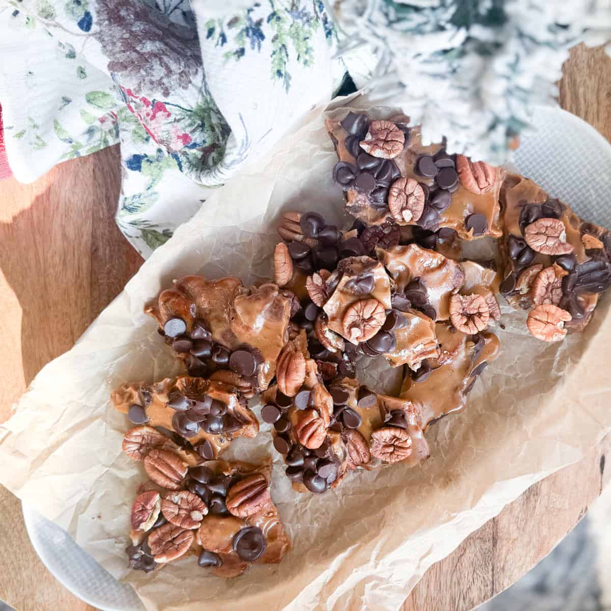 Parchment paper on a plate holds pieces of chocolate and pecan bark, topped with whole pecans and chocolate chips. A floral-patterned cloth and a snowy artificial tree are partially visible nearby.