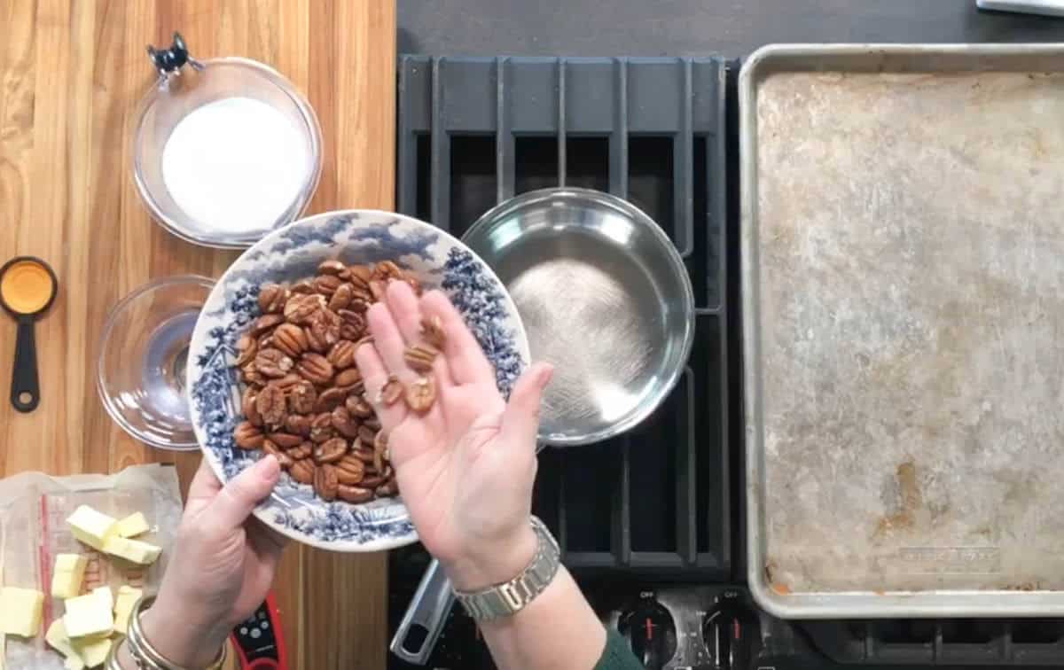 A person holds a bowl of pecans in one hand and a few pecans in the other over a stovetop. Nearby are a saucepan, a measuring cup of sugar, a stick of butter, a measuring spoon, and a baking sheet.