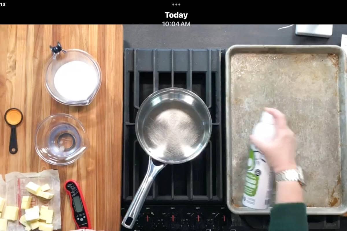Overhead view of a kitchen workspace with a saucepan on a stove, a wooden cutting board with ingredients including butter and sugar, and a hand spraying cooking spray onto a baking sheet.