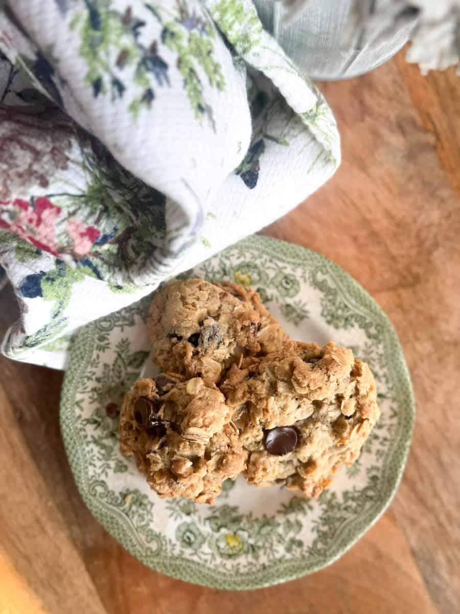 A green and white floral-patterned plate holds two chunky oatmeal cookies with chocolate chips. The plate sits on a wooden surface next to a folded floral-patterned cloth.