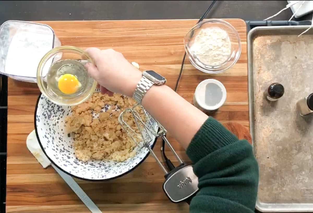 A person wearing a watch holds a small bowl with a raw egg above a larger bowl containing dough mixture on a wooden surface, surrounded by baking ingredients and utensils, including flour, vanilla, and a hand mixer.