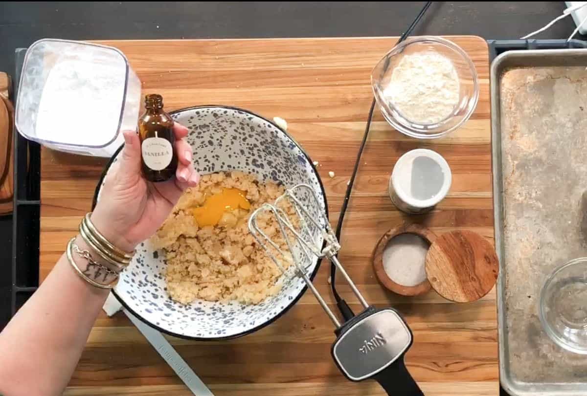 A hand holds a bottle of vanilla extract above a mixing bowl containing sugar, butter, and an egg. An electric hand mixer rests in the bowl. Measuring cups, flour, and a baking tray are on the wooden countertop.