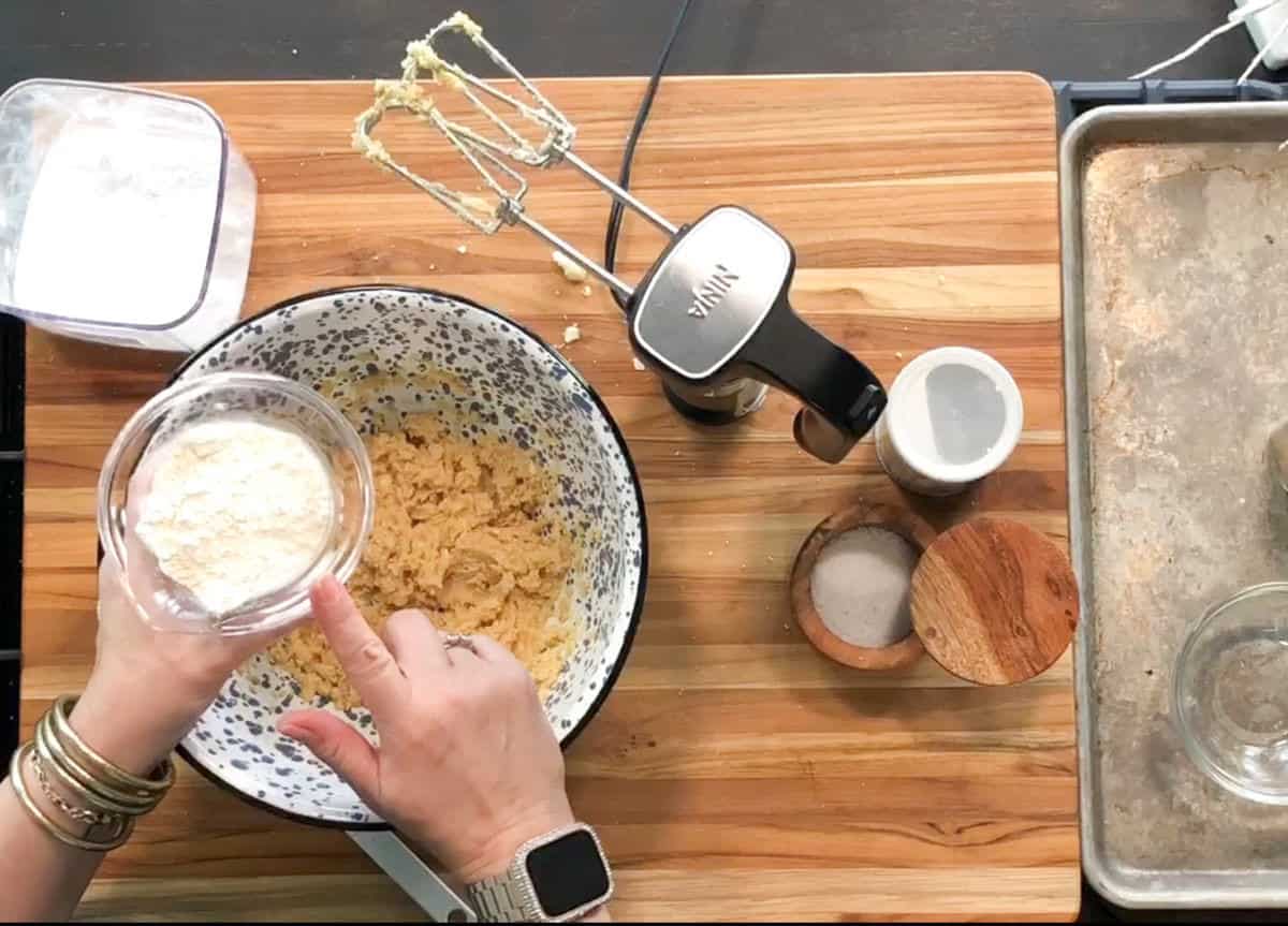 A person wearing bracelets and a smartwatch is preparing dough in a mixing bowl with an electric mixer. They are holding a small bowl of flour above the dough, ready to add it. Baking ingredients are on a wooden surface.