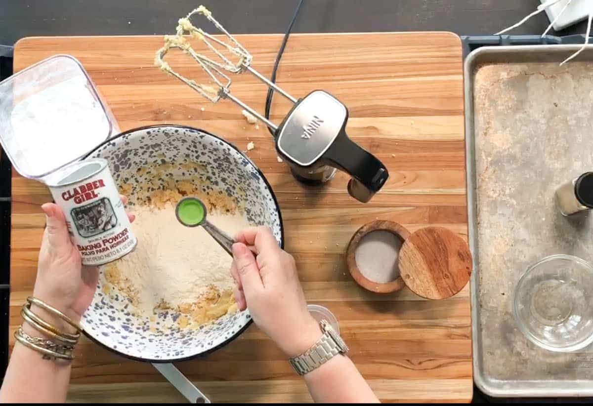 A person measures out baking powder from a Clabber Girl container into a mixing bowl with flour and batter. An electric mixer, salt cellar, and baking sheet are on the wooden countertop.