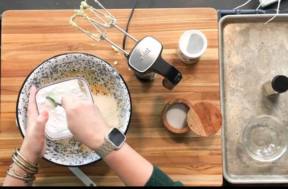 A person wearing a watch and bracelets measures powdered sugar over a mixing bowl on a wooden cutting board, with an electric mixer, spices, and a salt container nearby. A baking tray and small bowl are off to the side.