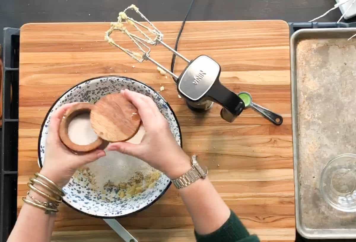 A person holding a wooden salt cellar adds an ingredient to a mixing bowl on a wooden cutting board, with a hand mixer and a metal baking tray nearby.
