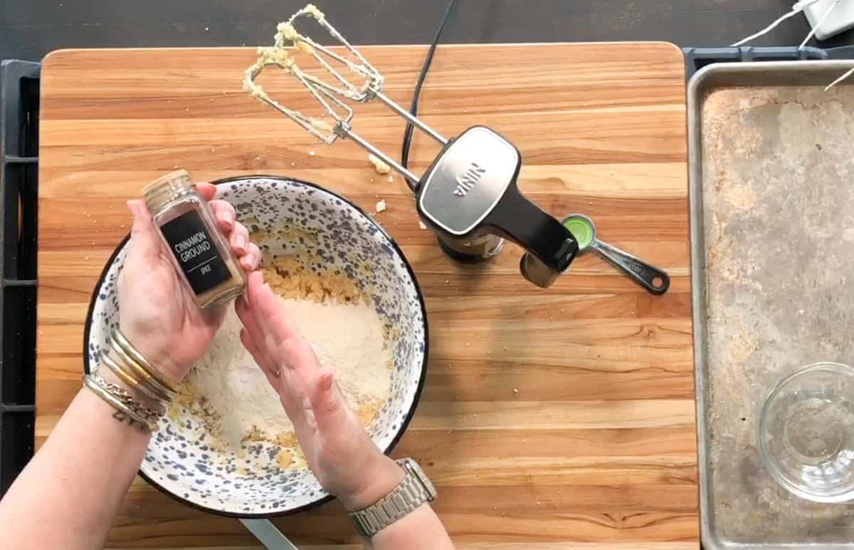A person holds a small jar of ground cinnamon over a large speckled mixing bowl filled with flour and other ingredients. A hand mixer, a measuring spoon, and a baking sheet are on the wooden surface nearby.